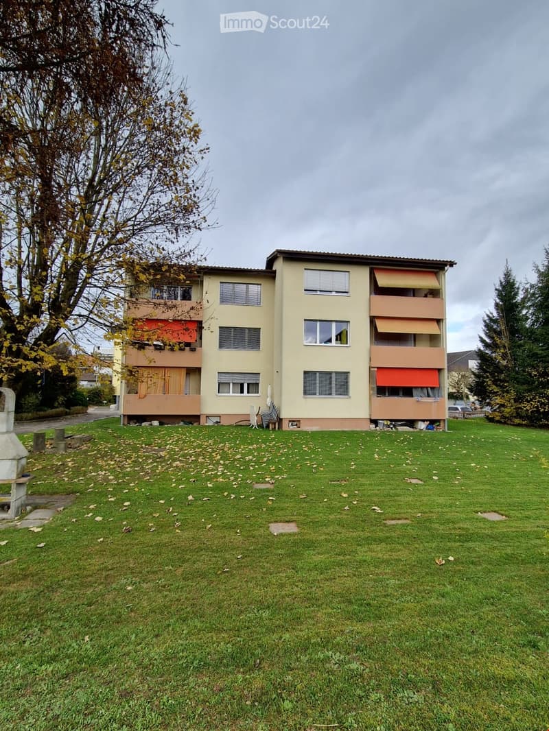 A three-story residential building with beige walls, multiple windows, and balconies with orange awnings. The building is surrounded by a grassy lawn with scattered leaves and trees.