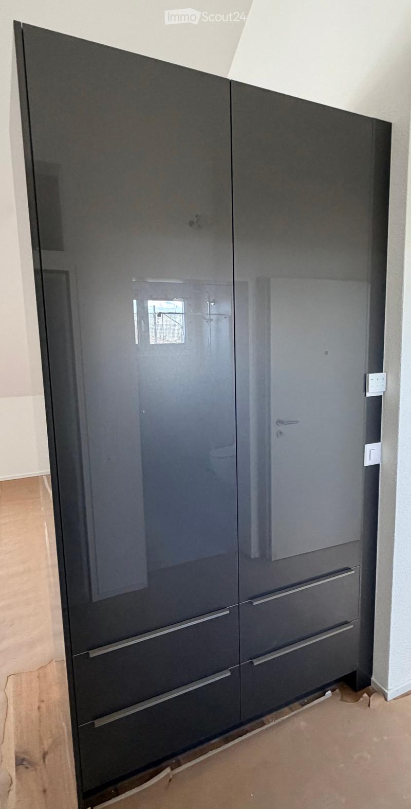 Empty room with a large, dark-colored wardrobe with mirrored doors and silver handles. The floor is light brown and the walls are white.