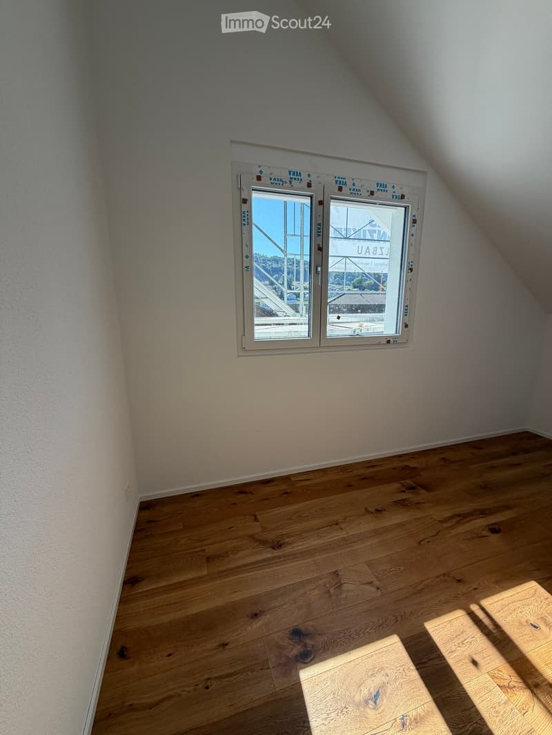 Empty attic room with wooden flooring and a large window offering a view of the outside. The walls are plain and the space is well-lit with natural light.