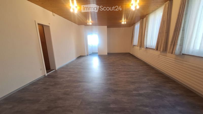 Empty room with wooden flooring, white walls, and large windows with curtains. The space is well-lit with ceiling lights and has a door leading to another area.