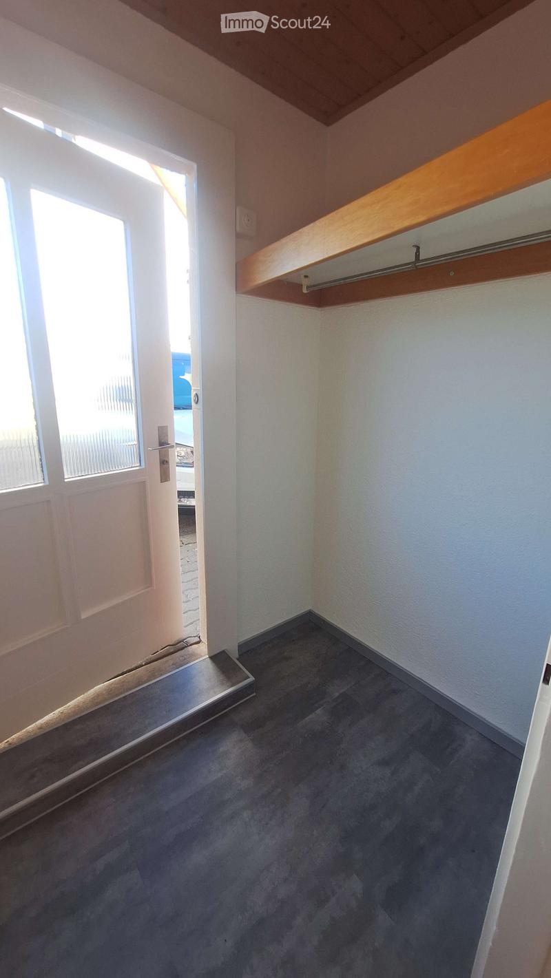 Empty room with grey carpeted floor, white walls, wooden shelf, and a door leading outside.