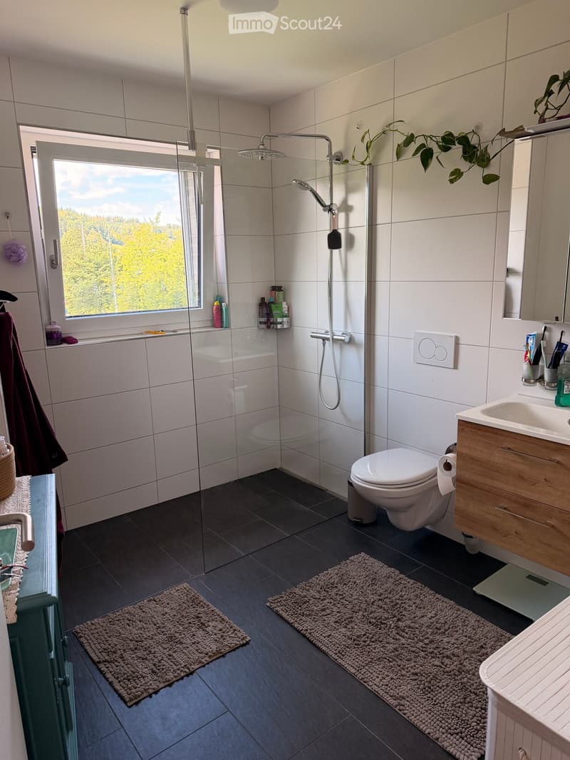 Modern bathroom with dark tiled floor, white tiled walls, a shower with glass enclosure, a toilet, a sink with wooden cabinet, and a window with a view of trees.