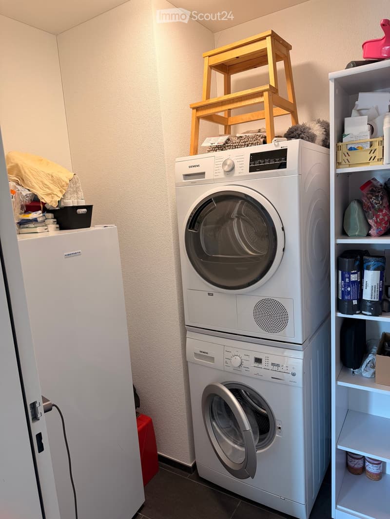 A small bathroom with a washing machine, dryer, refrigerator, shelf, and a wooden stool.