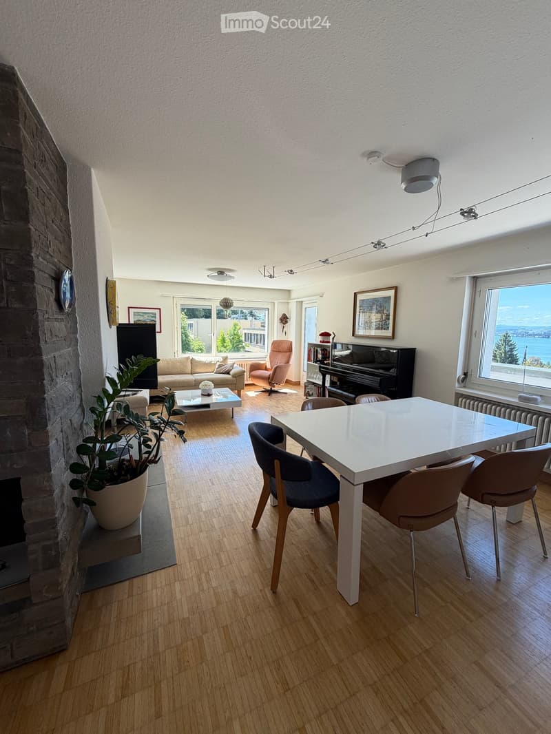 Open-plan living and dining area with wooden flooring, large windows offering a scenic view, a sofa, coffee table, dining table with chairs, a piano, and a potted plant.