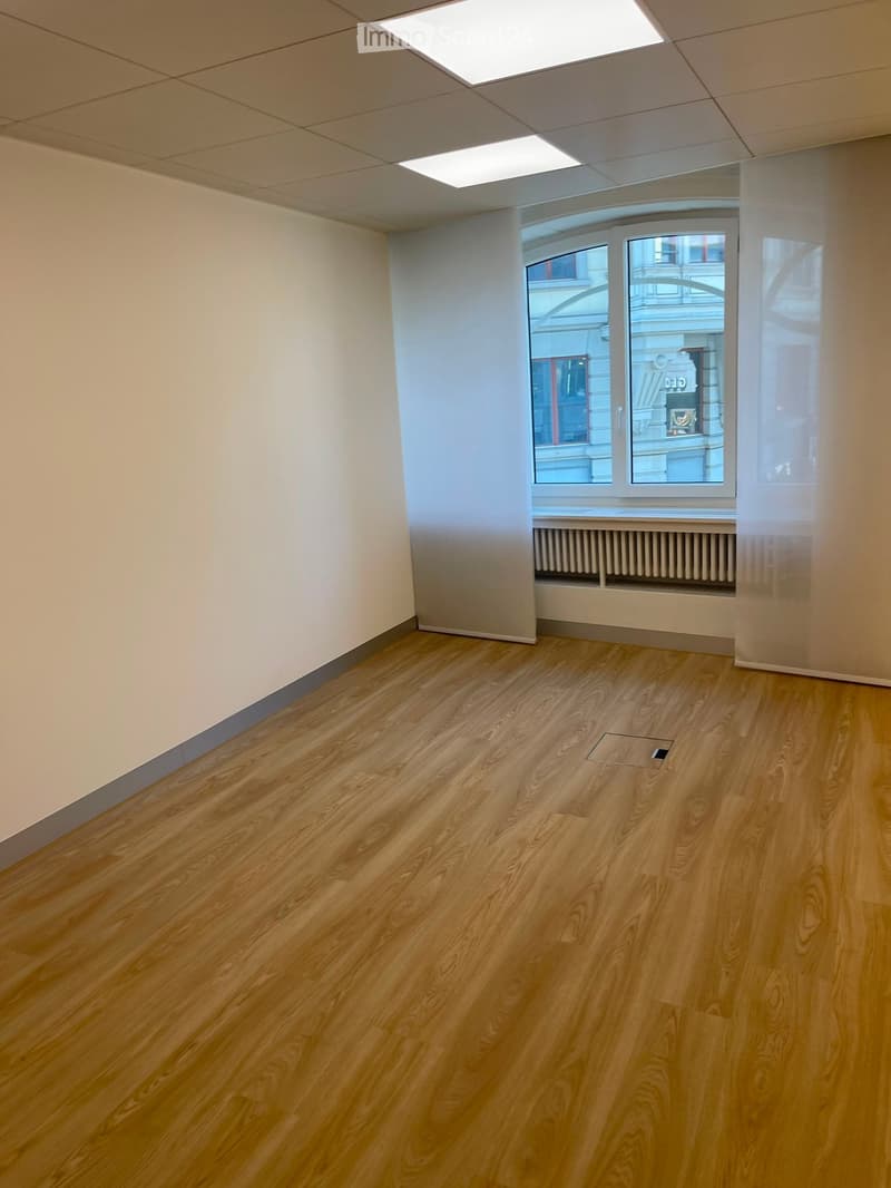 Empty room with light wooden flooring, white walls, and large windows with a view of the street. The ceiling has recessed lighting and a skylight.