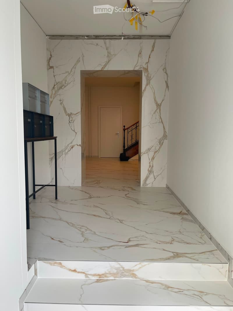 Elegant hallway with marble flooring and walls, featuring a staircase with a black railing and a small table with a blue cabinet on the left side.