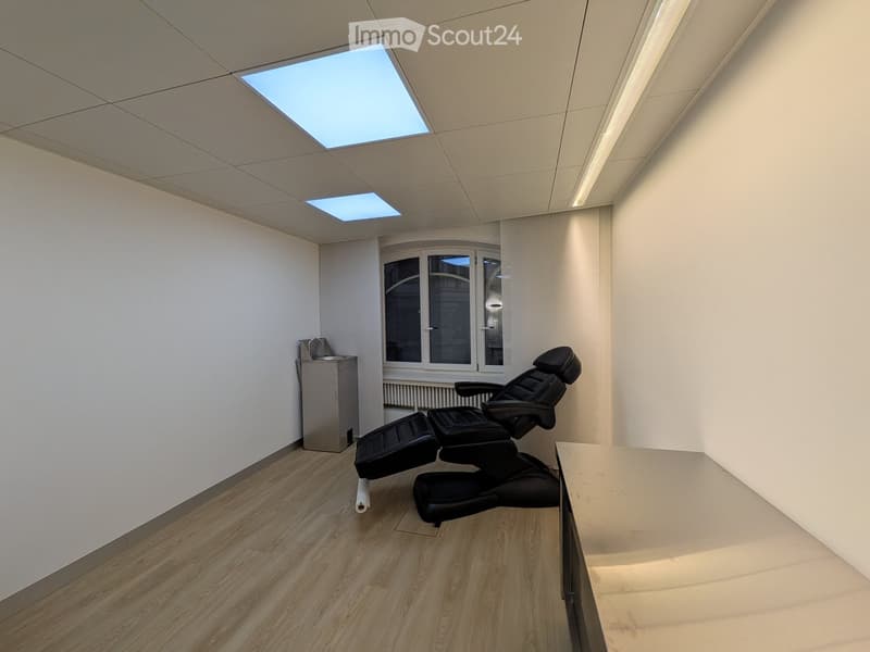 Modern, minimalist room with wooden flooring, white walls, and a large window. Features a black massage chair and a small sink unit.