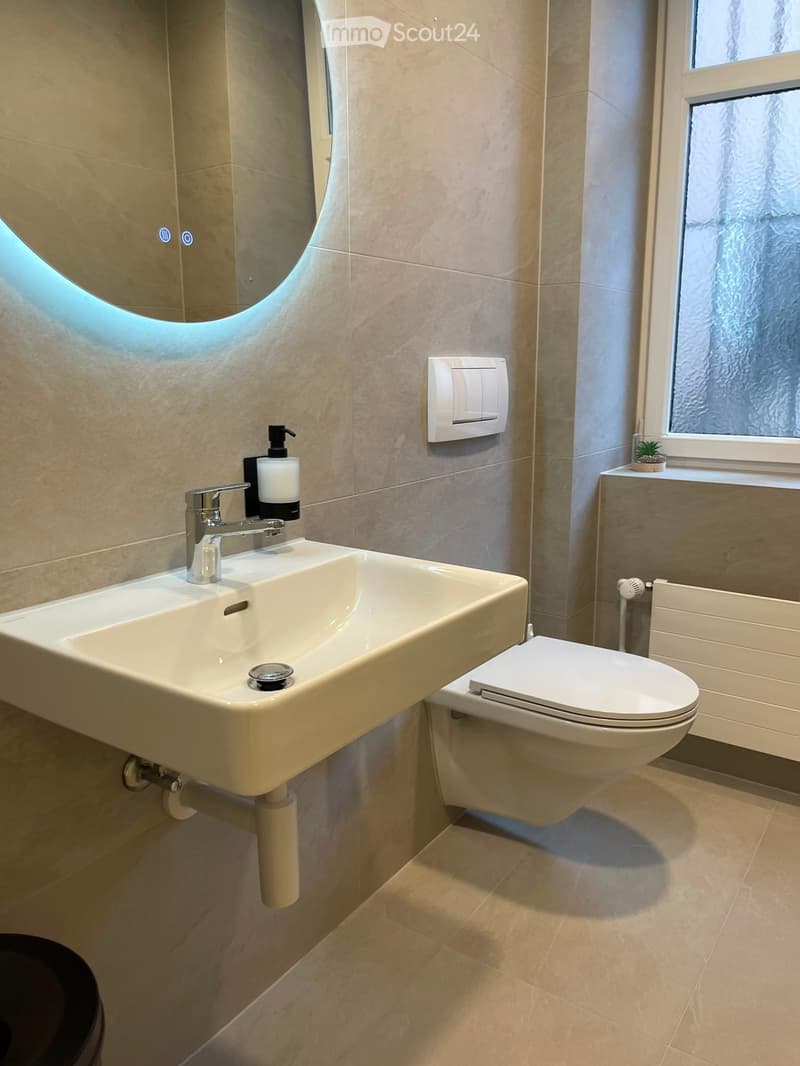 Modern bathroom with a white sink, chrome faucet, toilet, large mirror with LED lighting, and a small window with frosted glass. Light beige tiles cover the walls and floor.