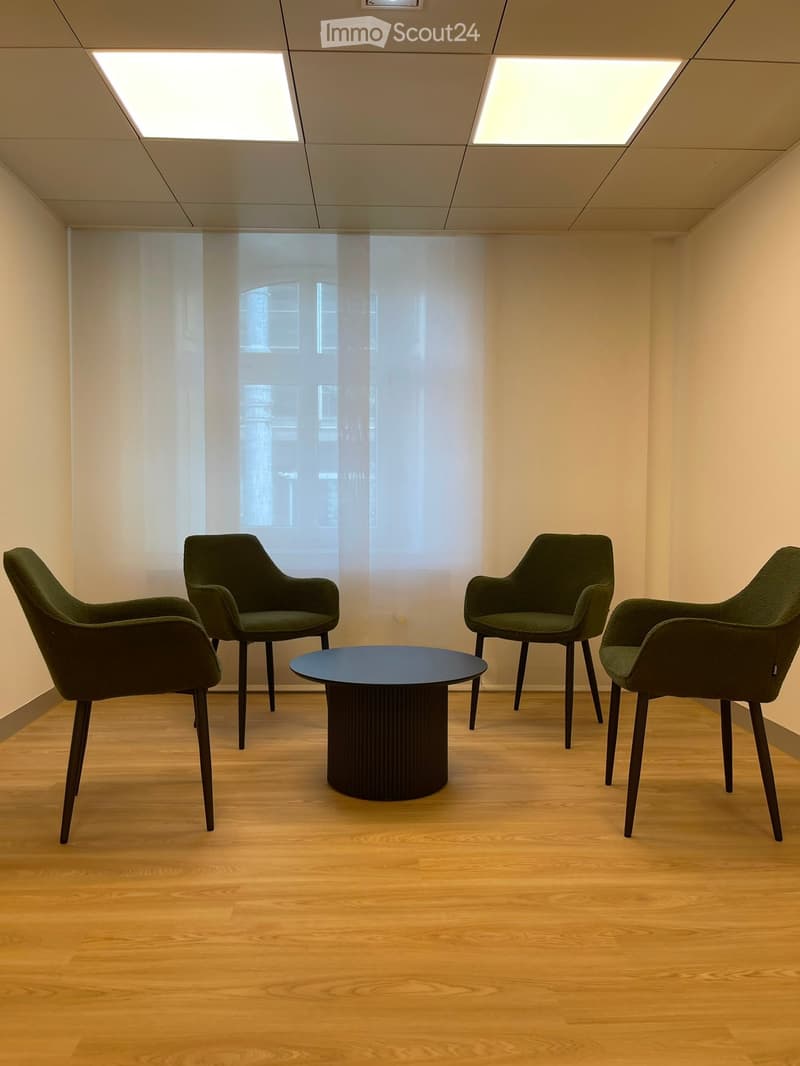 A modern, minimalist room with light wooden flooring, four dark green chairs, and a round black coffee table. The room features large windows with sheer curtains and recessed ceiling lighting.
