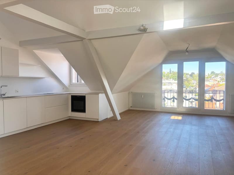 Bright, empty attic space with wooden flooring, white walls, and large windows offering a view of the city. The room features built-in white kitchen cabinets and a sink, but no furniture or appliances are present.