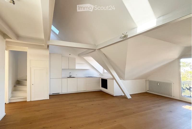 Bright, airy attic space with wooden flooring, white cabinets, sink, and oven. Slanted ceilings with skylights and large windows provide natural light.