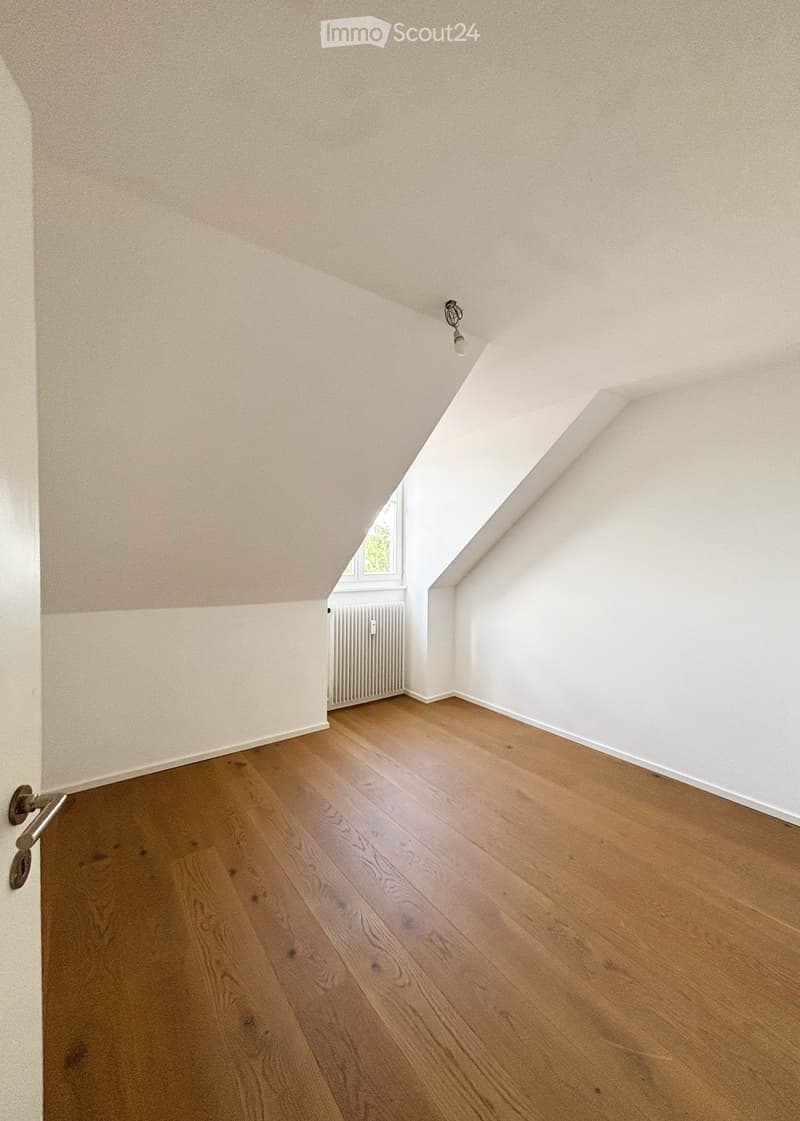 Empty attic room with wooden flooring, white walls, and a small window letting in natural light. The ceiling has a sloped design typical of attic spaces.