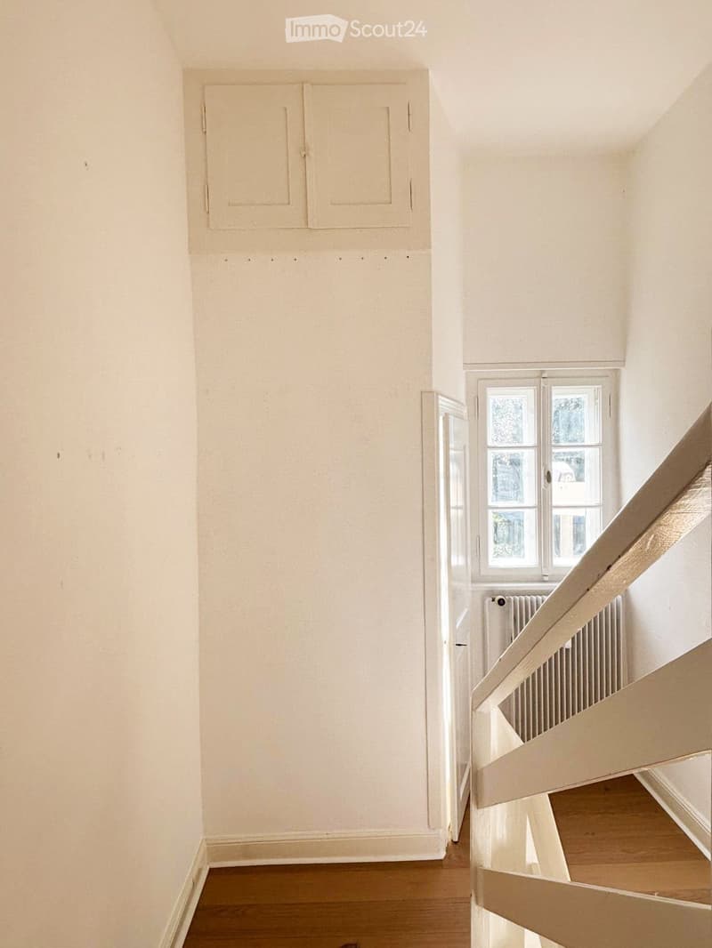 Empty hallway with light-colored walls, wooden flooring, and a staircase leading upwards. A window with white frames is visible on the right side, and a radiator is placed beneath it.