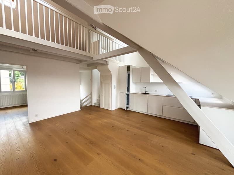 Open-plan space with wooden flooring, white walls, and a small kitchen area featuring cabinets and a sink. The room has a staircase leading to an upper level with a balcony overlooking the space.