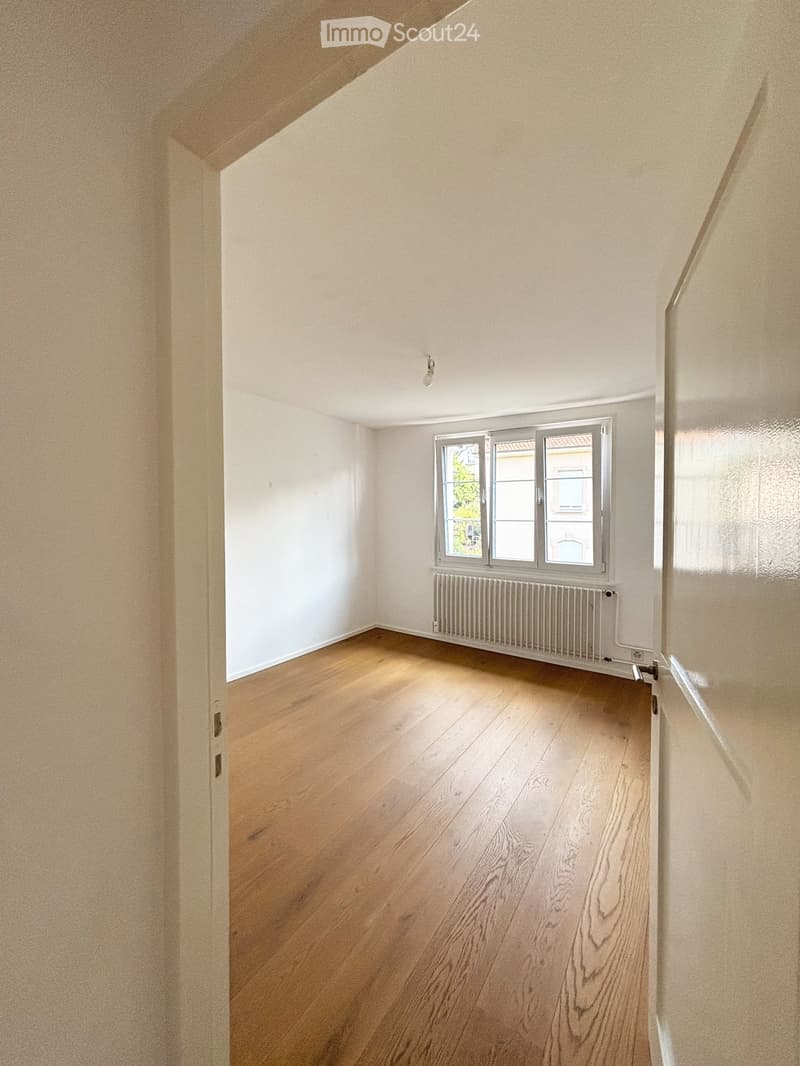 Empty room with wooden flooring, white walls, a window with a radiator below, and a door visible on the left side.