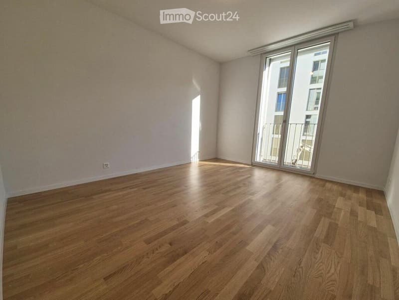 Empty room with wooden flooring and large sliding glass doors leading to a balcony. Light-colored walls and natural light from the windows.