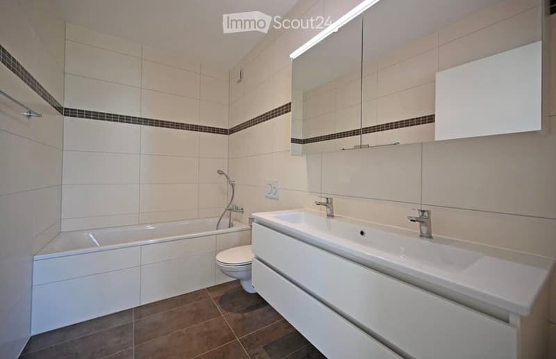 Modern bathroom with white tiles, a bathtub, toilet, double sink, and a large mirror with built-in lighting.