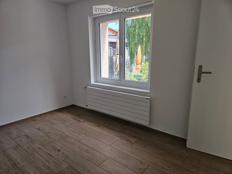 Empty room with wooden flooring, white walls, a radiator, and a large window with a view of a garden.