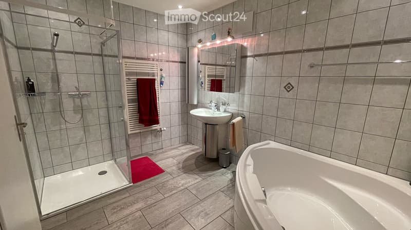 Modern bathroom with a bathtub, sink, mirror, and glass-enclosed shower. Features tiled walls and floor, a towel rack with a red towel, and a small red bath mat in the shower area.