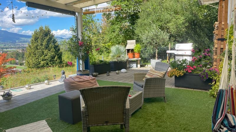 Well-maintained patio with artificial grass, wicker chairs, a bean bag, and a hanging hammock. Features a grill, potted plants, and a scenic mountain view.