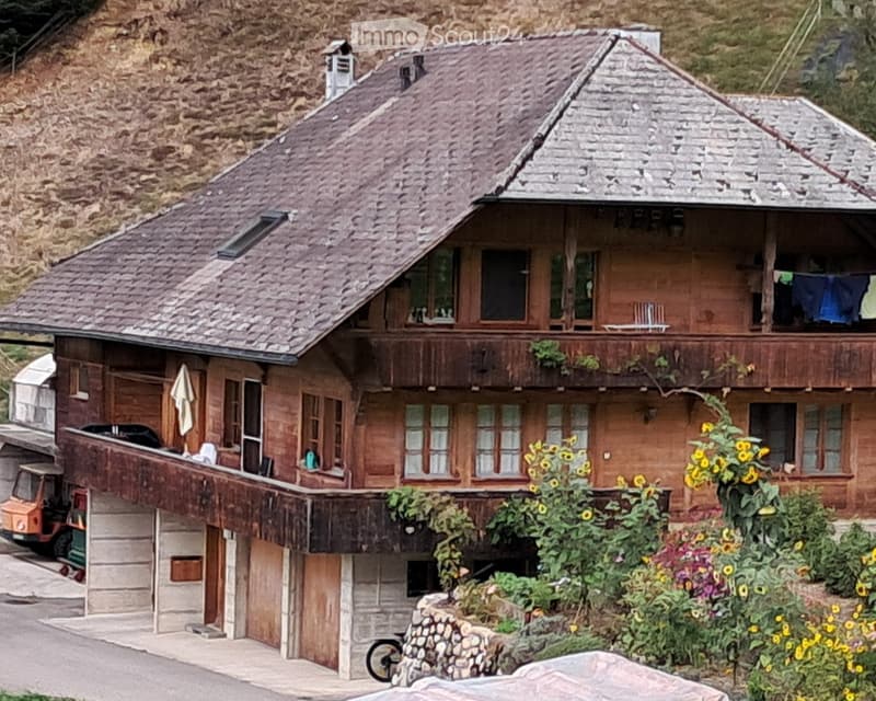 A two-story wooden house with a stone base, large windows, and a balcony. The roof is covered with brown tiles, and the house is surrounded by a garden with sunflowers and other plants. A small vehicle is parked near the garage.