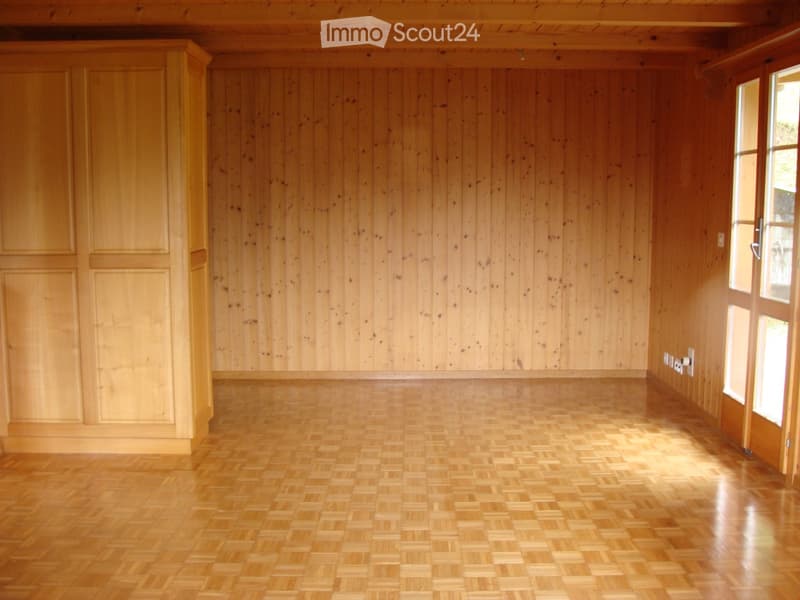 Empty room with wooden walls and herringbone-patterned wooden flooring, featuring a large window and built-in cabinetry.