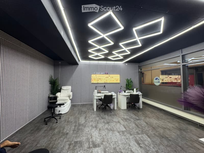 Modern, well-lit salon or spa room with grey carpet, white walls, and large windows. Features a pedicure chair, two desks with office chairs, a small plant, and decorative lighting.