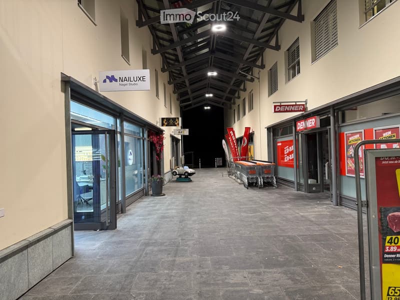 A modern indoor corridor with glass storefronts, shopping carts, and signage for stores like "NAILUXE" and "DENNER".