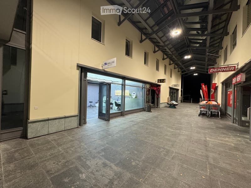 Wide, open corridor with tiled flooring, glass storefronts, and visible signage for businesses like 'NAIL LUXE' and 'DENNER'.