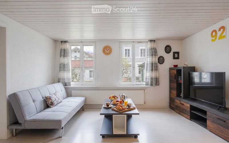 Bright living room with white walls, large windows, grey sofa, coffee table with breakfast items, television, and decorative wall clocks.