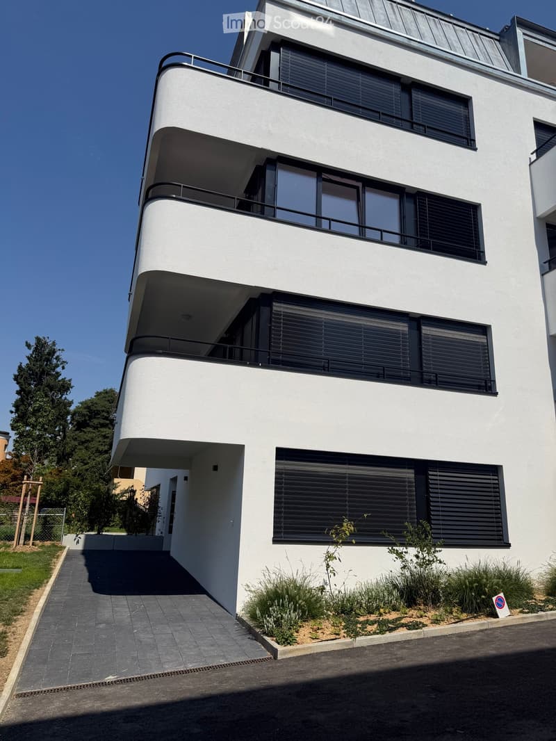 Modern white building with multiple levels, large windows, and balconies. Features contemporary design with clean lines and a raised ground floor.