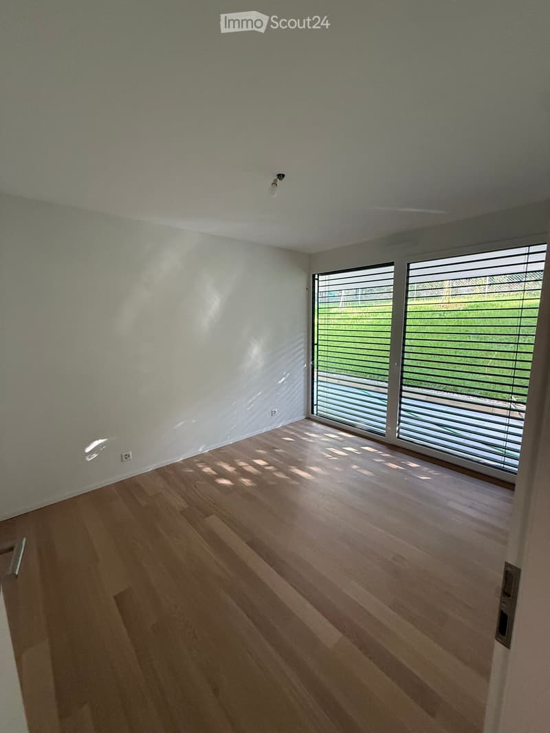 Empty room with light-colored wooden flooring and white walls, featuring large windows with blinds and a ceiling light fixture.