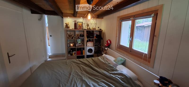 Cozy bedroom with a bed, wooden shelf, hanging lamp, teddy bear, hat, and various decorative items. The room has wooden beams on the ceiling and large windows letting in natural light.