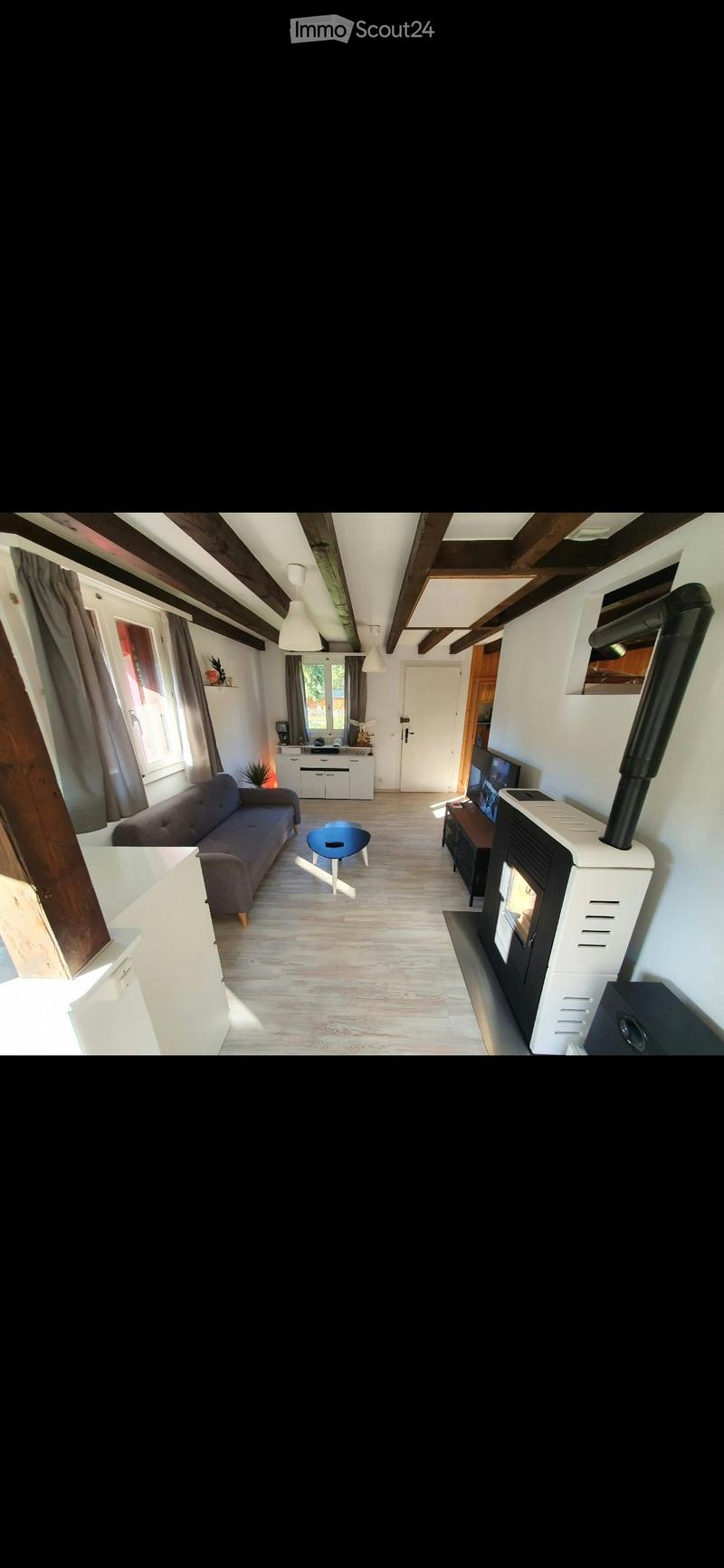 Bright living room with wooden flooring, exposed wooden beams, large windows with curtains, a sofa, coffee table, TV, fireplace, and a small table with a chair.
