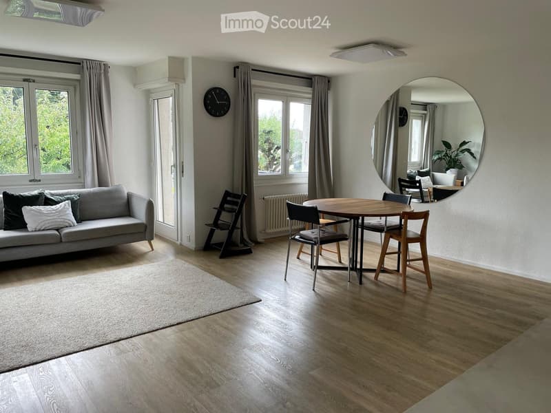 Open-plan living and dining area with wooden flooring, a grey sofa, a round wooden dining table with four chairs, a large round mirror, and a clock on the wall.