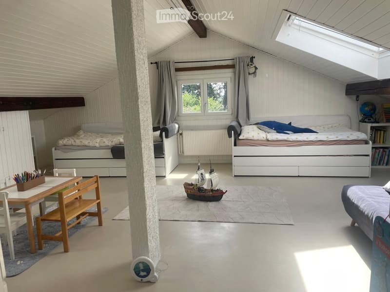 Bright, airy bedroom with two single beds, a small table and chairs, a toy ship on the floor, and a bookshelf with books and a globe. The room has white wooden paneling and a slanted ceiling with skylights.