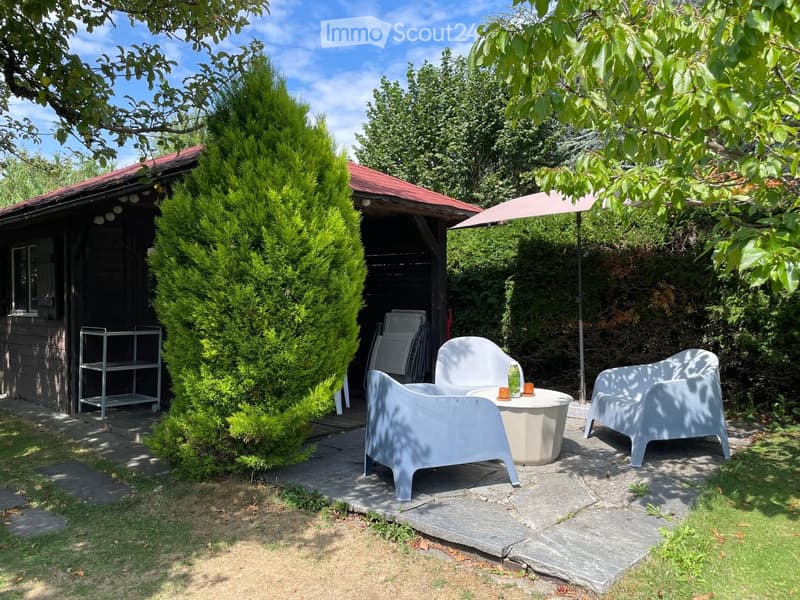 Outdoor patio area with white chairs, a small table, and an umbrella, surrounded by greenery and a wooden shed.