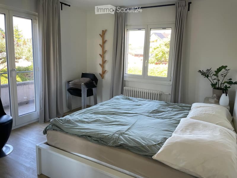 Bright bedroom with a double bed, light blue duvet, white pillows, a small table with a plant, a chair, and a radiator. Large windows offer a view of greenery outside.