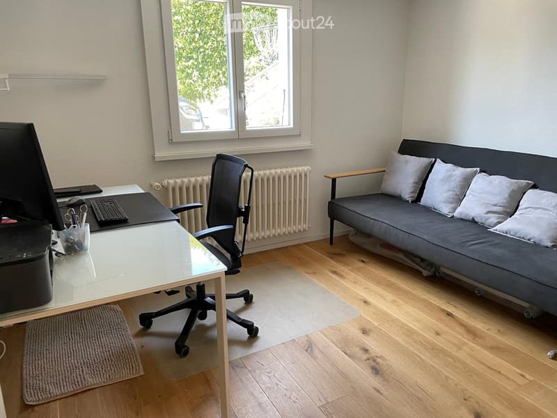 A modern, multi-functional room with a grey sofa, a black office chair, a white desk, and a radiator. The room features large windows with a view of greenery outside.