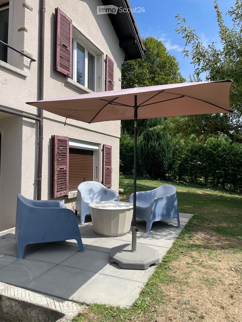 Outdoor patio area with a large umbrella, three chairs, and a round table, adjacent to a house with wooden shutters.