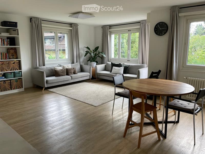 Bright living room with wooden flooring, two grey sofas, a round wooden dining table with chairs, a bookshelf, and a potted plant near the window.