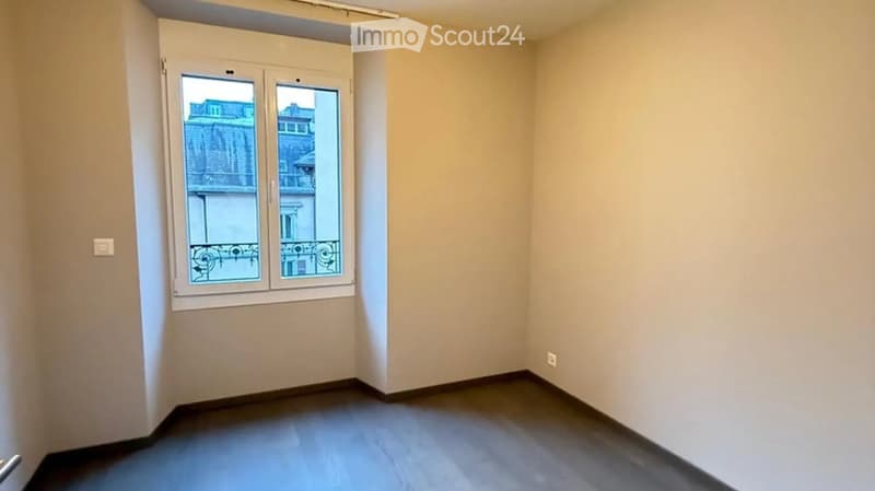 Empty room with light-colored walls, dark flooring, and a large window with a view of a building and balcony.