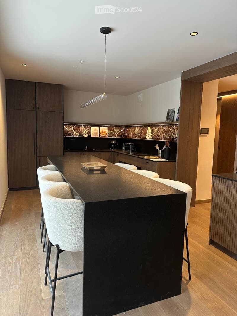 Modern kitchen with a black kitchen island, white stools, wooden flooring, and sleek cabinetry.