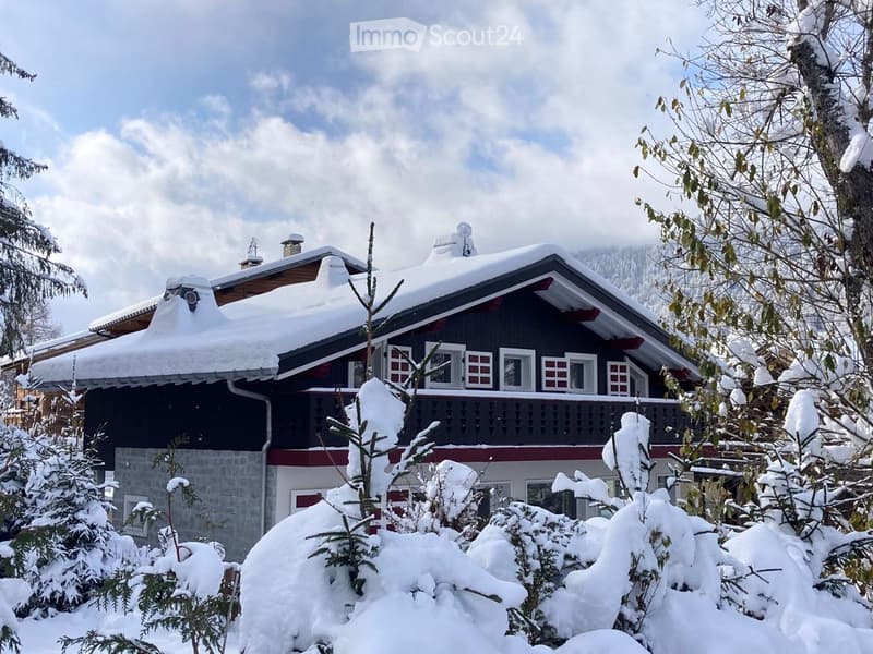 A two-story house with a snow-covered roof and balcony, surrounded by snow-covered trees and shrubs.