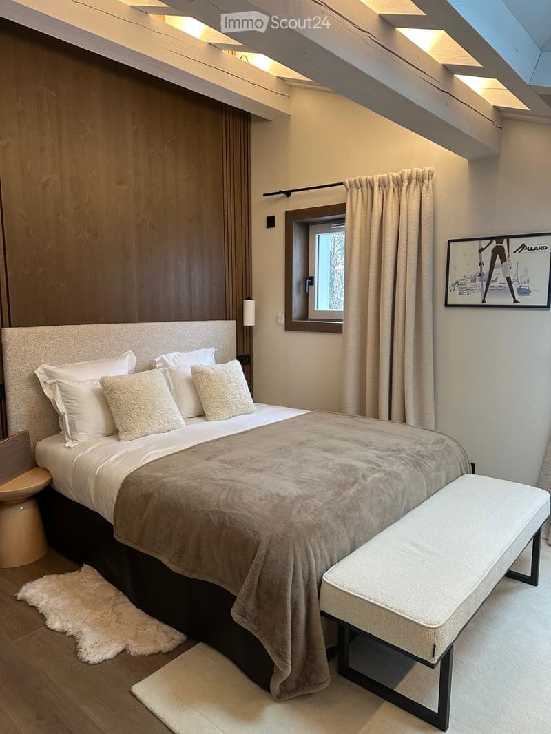 Modern bedroom with a large bed, bench, stool, and artwork on the wall. The room features wooden beams and a skylight.