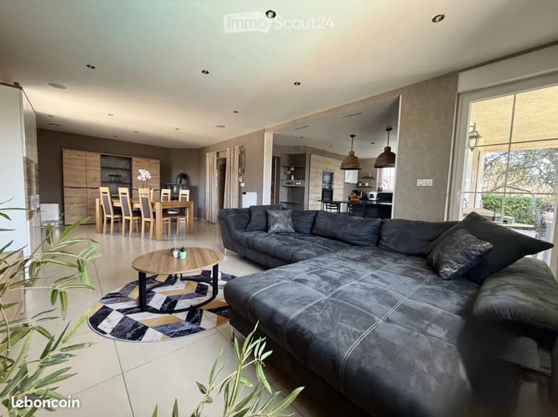 Modern living and dining area with a grey sectional sofa, wooden coffee table, dining table with chairs, and kitchen area with cabinets and pendant lights.
