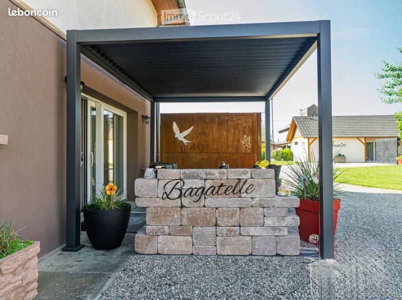 A covered outdoor area with a stone sign that reads "Bagatelle" and several potted plants, including a large black pot with a yellow flower. The area is connected to a house with a wooden door featuring a decorative bird and butterfly design.