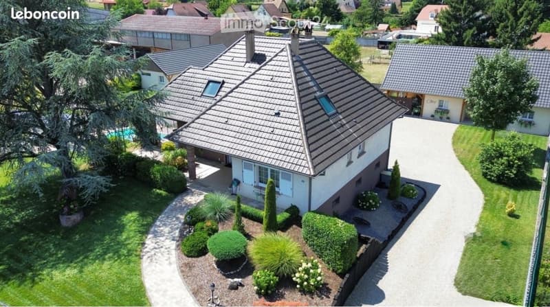 Aerial view of a two-story house with a gray tiled roof, surrounded by a well-maintained garden with shrubs and trees. A swimming pool is visible in the background, along with neighboring houses and a driveway leading to the property.
