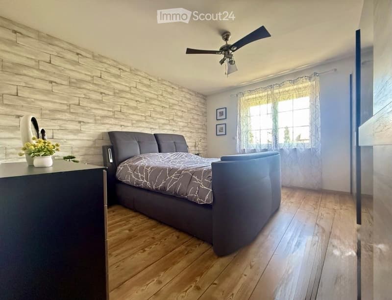 Modern bedroom with wooden flooring, dark grey bed, nightstand, flower pot, and ceiling fan.