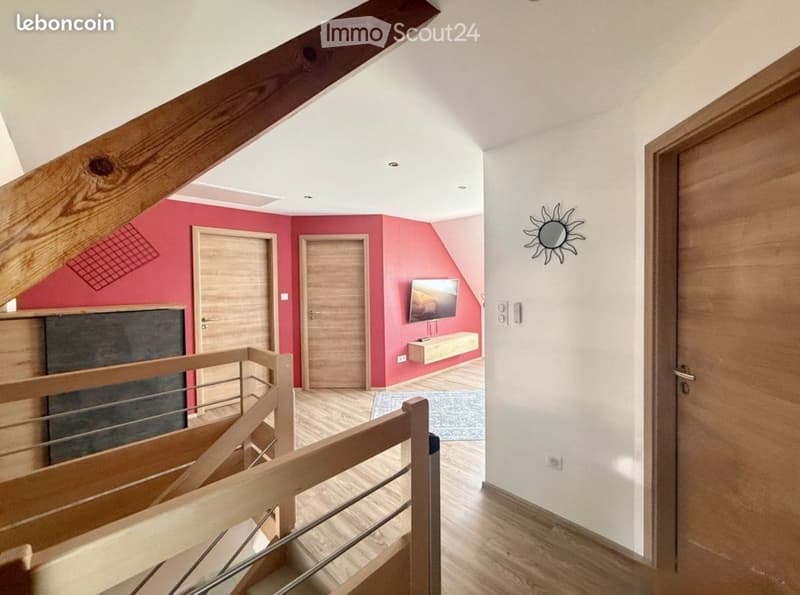Modern hallway with wooden flooring, white walls, and a staircase leading upwards. Features include a TV mounted on a pink accent wall, a small wooden shelf, and a sun-shaped wall decoration.